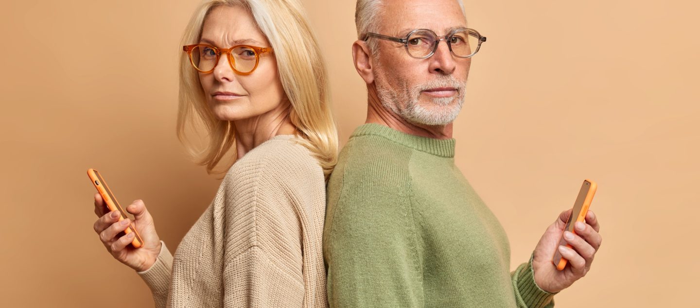 serious-senior-woman-her-husband-hold-modern-gadgets-read-media-spend-free-time-internet-ignore-each-other-stand-backs-wear-spectacles-sweater-isolated-beige-wall