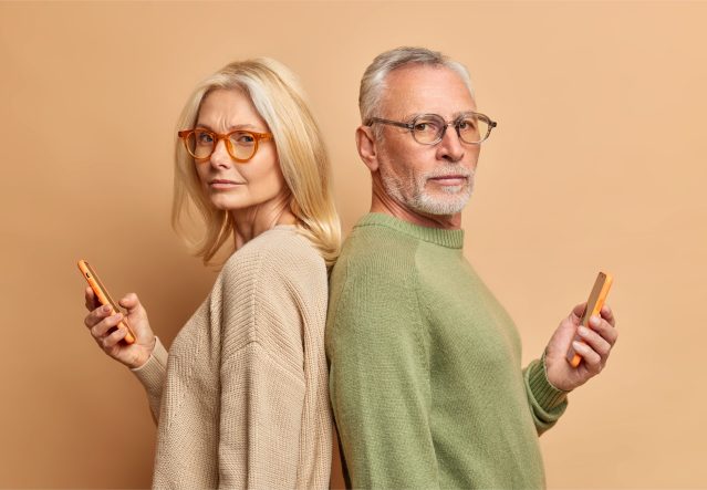 serious-senior-woman-her-husband-hold-modern-gadgets-read-media-spend-free-time-internet-ignore-each-other-stand-backs-wear-spectacles-sweater-isolated-beige-wall